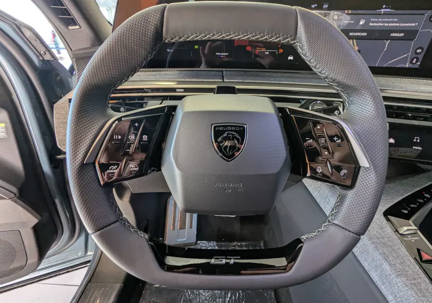Vue rapprochée du volant cuir perforé noir avec logo Peugeot et badge GT du Peugeot 3008 Bleu Ingaro 2026.
