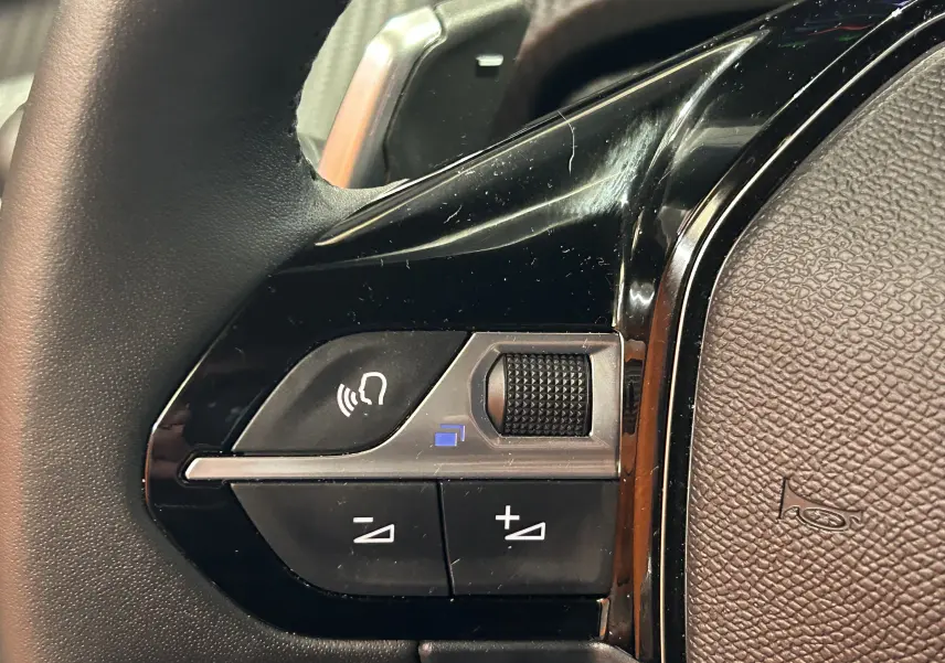 Gros plan sur les commandes noires du volant du Peugeot 2008 II Ph.2 Allure Hybrid 145, détails de la molette et boutons.
