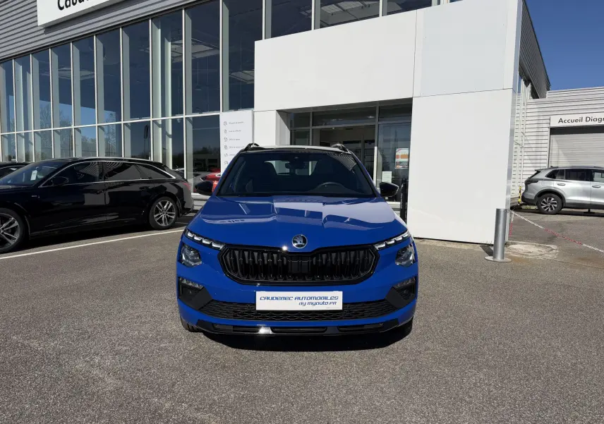 Vue de face d'un Skoda Kamiq bleu 2025 avec calandre noire et projecteurs LED haute gamme, stationné devant un bâtiment.