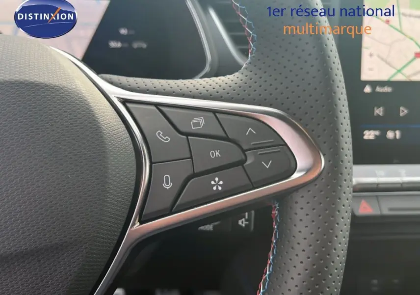 Vue rapprochée du volant en cuir perforé avec commandes multifonctions, tableau de bord et écran tactile en arrière-plan.