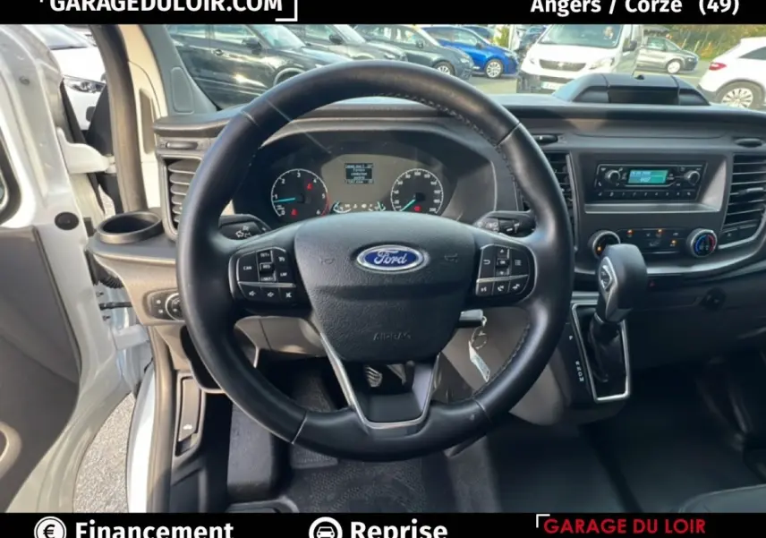 Vue intérieure centrée sur le volant multifonctions en cuir du Ford Transit Custom blanc, avec tableau de bord et levier auto visibles.