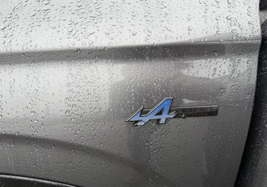 Gros plan sur l'écusson Esprit Alpine bleu sur la carrosserie gris schiste mouillée du Renault Austral 2025.