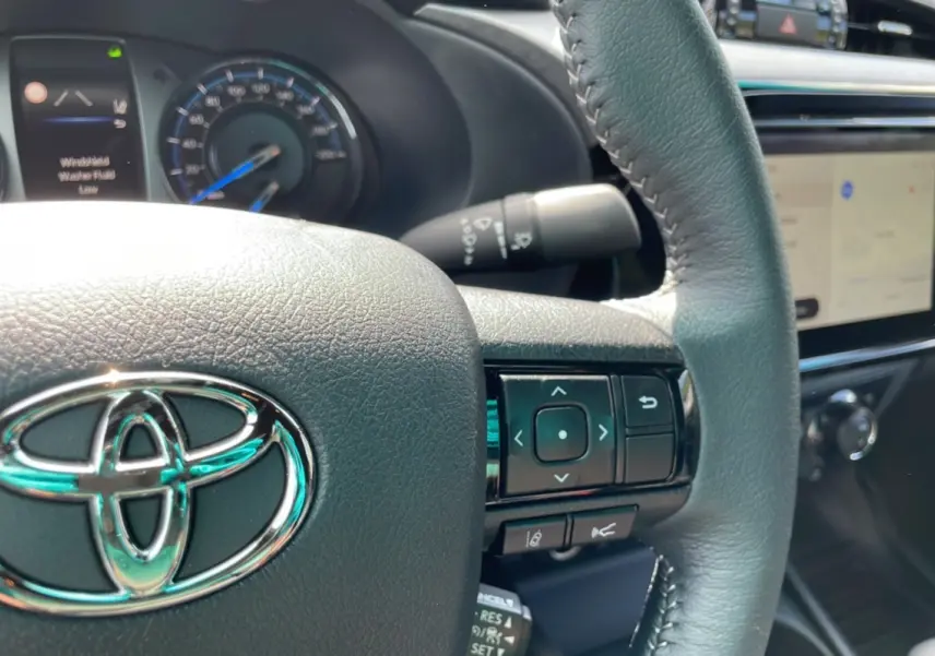 Gros plan sur le volant en cuir noir du Toyota Hilux 2025, avec commandes intégrées et tableau de bord numérique visible.