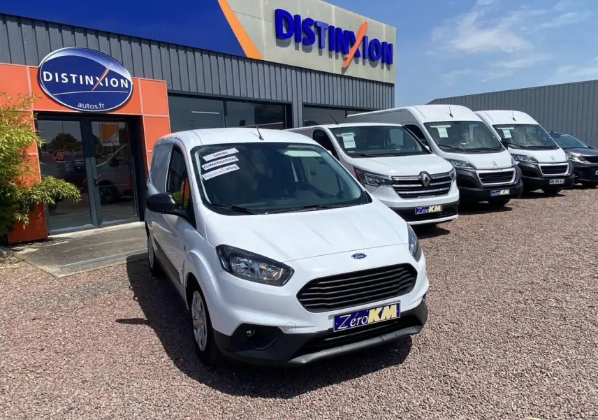 Vue 3/4 avant d'un Ford Tourneo Courier blanc stationné devant un concessionnaire, avec calandre noire et enjoliveurs visibles.