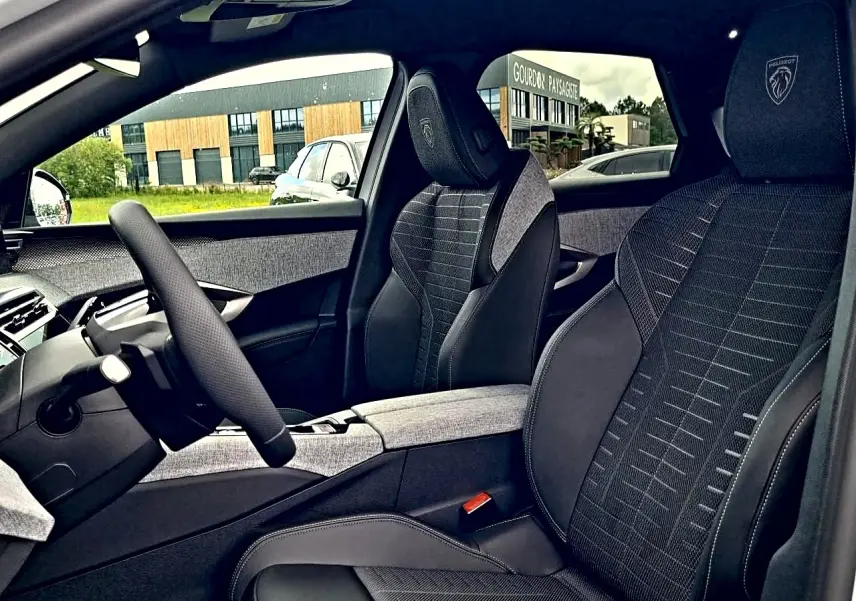 Intérieur avant droit du Peugeot 3008 2026, sièges tissu noir avec logo Peugeot brodé sur appuie-tête.