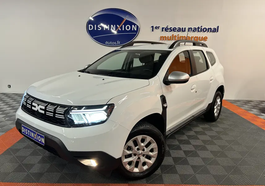 Vue 3/4 avant droite d'un Dacia Duster blanc 2023 avec feux allumés et barres de toit noires en intérieur showroom.