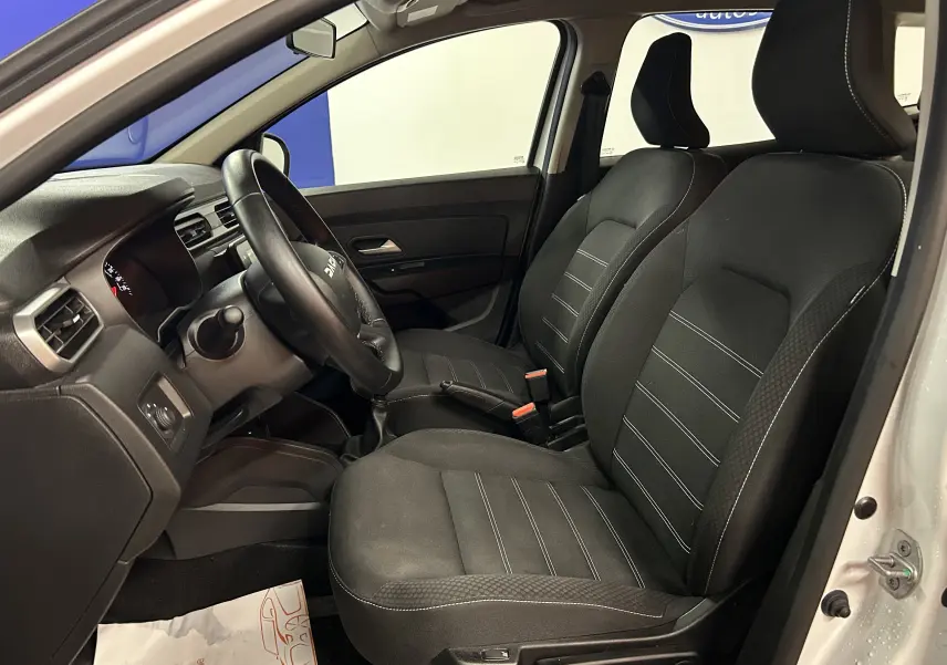 Vue intérieure côté conducteur du Dacia Duster blanc 2023, mettant en valeur les sièges chauffants en tissu noir avec surpiqûres blanches.