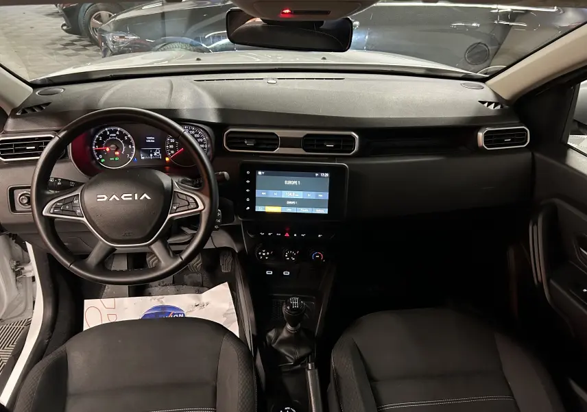 Vue intérieure avant du Dacia Duster blanc 2023, tableau de bord avec écran tactile et volant multifonction.