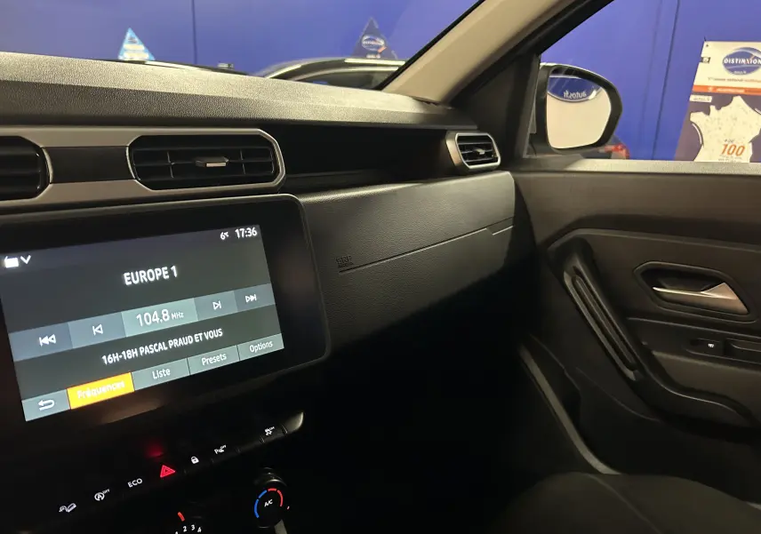 Vue intérieure côté passager du tableau de bord noir du Dacia Duster 2023 avec écran tactile et commandes climatisation.
