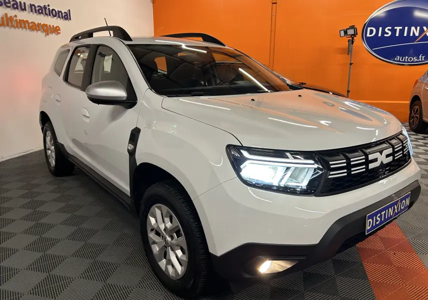 Vue 3/4 avant droite d'un Dacia Duster blanc 2023 avec phares LED allumés et barres de toit noires.