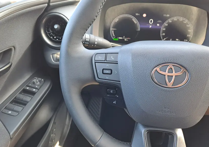 Gros plan sur le volant noir du Toyota C-HR gris minéral 2025, avec commandes intégrées et tableau de bord digital en arrière-plan.