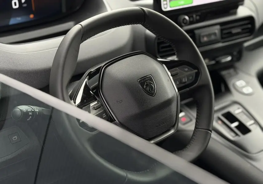 Vue rapprochée du volant cuir noir du Peugeot Rifter 2026 avec commandes intégrées et tableau de bord numérique partiel.