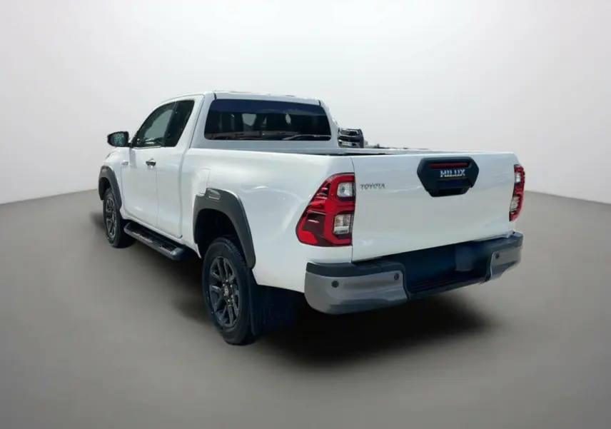 Toyota Hilux 4x4 blanc pur en 3/4 arrière droit, avec protections noires et jantes foncées visibles.