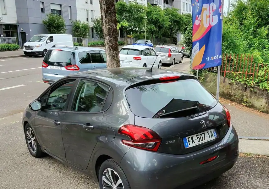 Vue 3/4 arrière droite d'une Peugeot 208 gris foncé métal, avec feux arrière LED et cinq portes visibles en milieu urbain.