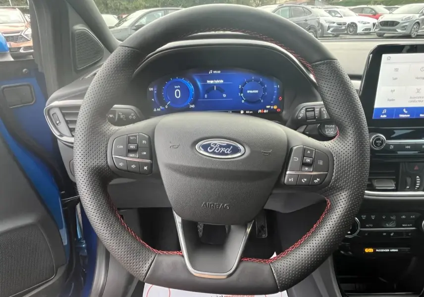 Vue intérieure centrée sur le volant perforé noir avec surpiqûres rouges d’un Ford Puma bleu, tableau de bord numérique visible.