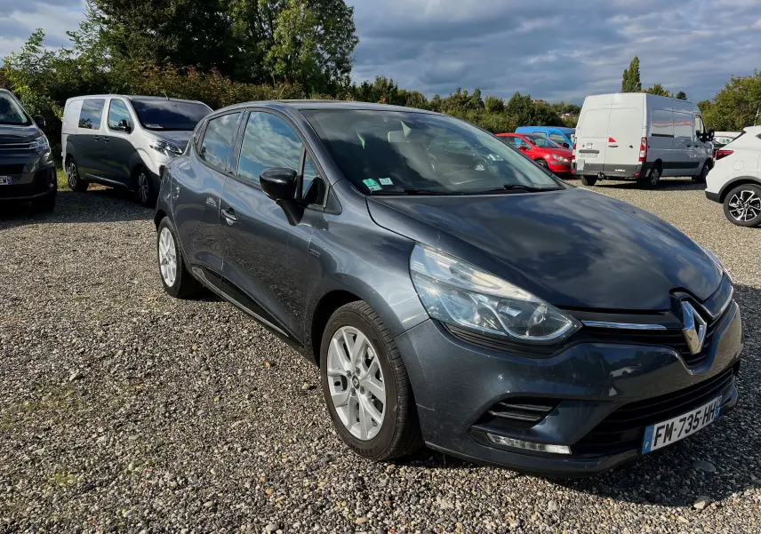 Renault Clio IV gris foncé vue en 3/4 avant droit sur un parking gravillonné par temps nuageux.