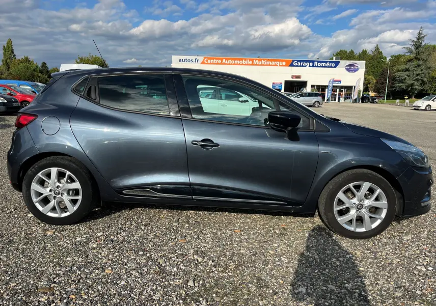 Profil côté gauche d'une Renault Clio IV gris foncé 2019 garée sur un sol gravillonné devant un garage multimarque.