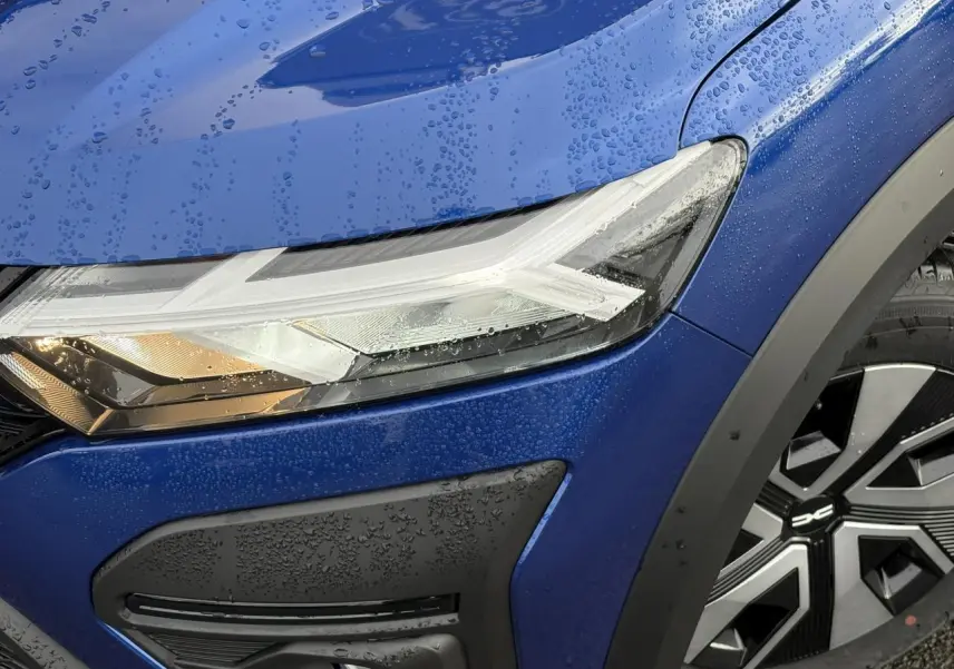 Gros plan sur le phare avant gauche et la roue avec enjoliveur 16" d'une Dacia Sandero Stepway Bleu Iron mouillée.