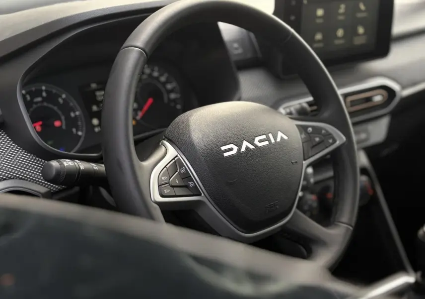 Vue rapprochée du volant noir Dacia avec commandes intégrées et tableau de bord numérique en arrière-plan.
