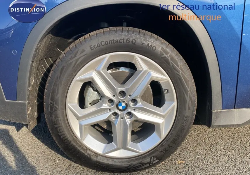 Gros plan sur la roue avant gauche d'un BMW X1 bleu phytonic metal avec jante argentée cinq branches.