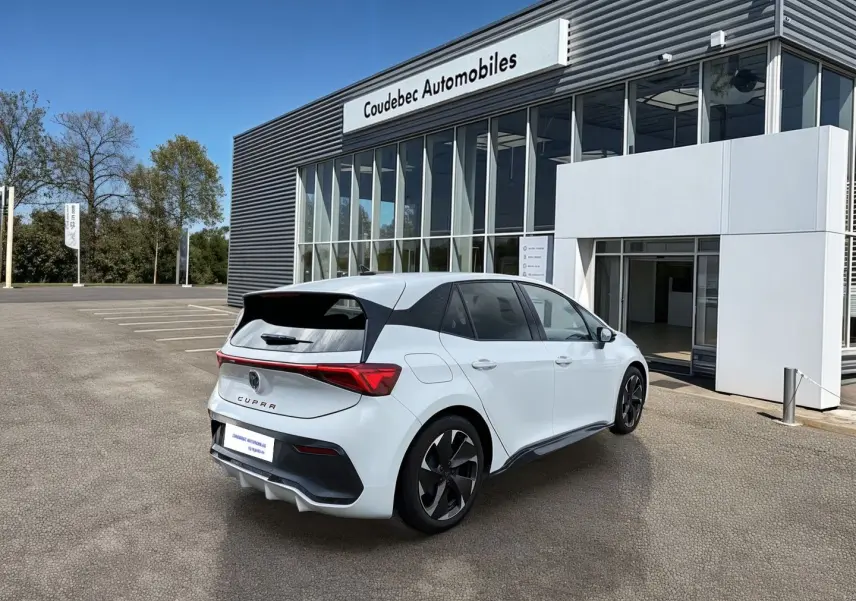 Vue 3/4 arrière droite d'une CUPRA Born blanche avec jantes noires Typhoon devant un bâtiment moderne.