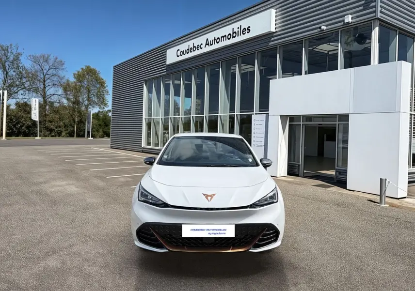 Vue avant d'une CUPRA Born électrique blanche avec logo cuivré, feux LED et calandre noire devant un bâtiment moderne.