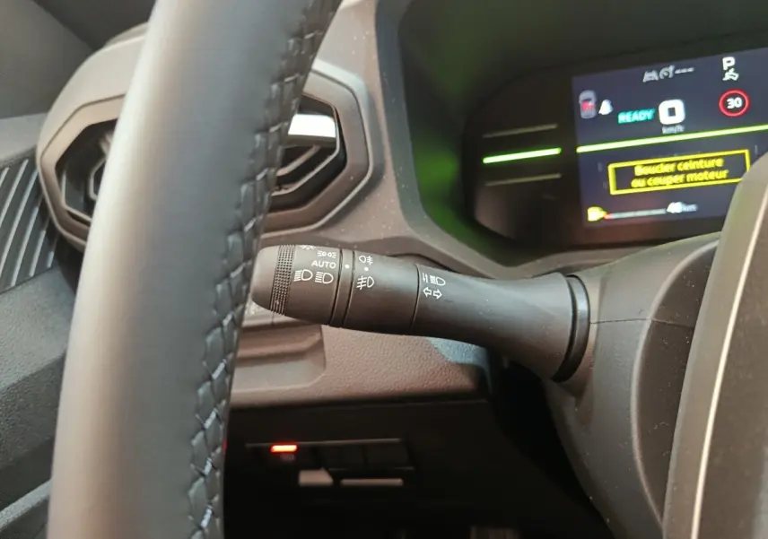 Gros plan sur la commande des phares à gauche du volant noir du Dacia Duster Journey Hybrid 155 avec tableau de bord numérique.