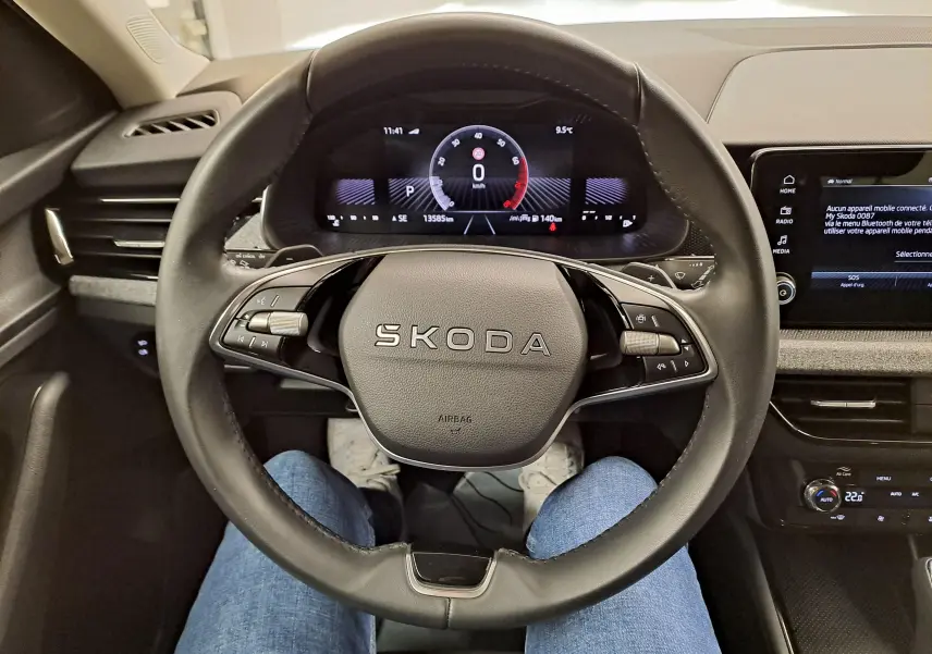 Vue intérieure centrée sur le volant SKODA d'une Scala 2025, avec tableau de bord numérique et commandes au volant visibles.