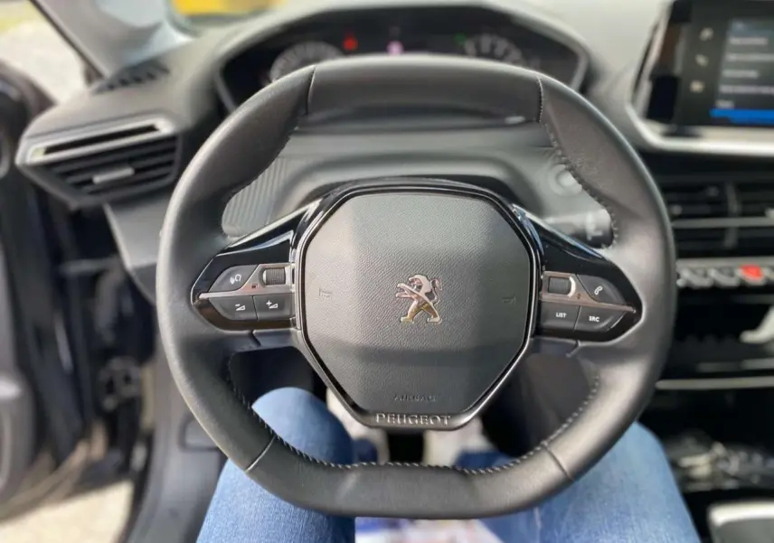 Vue rapprochée du volant noir de la Peugeot 208 2021 avec commandes intégrées et tableau de bord en arrière-plan.