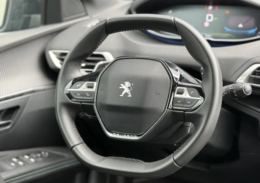 Volant compact cuir noir Mistral avec logo Peugeot au centre, tableau de bord et commandes intégrées visibles en arrière-plan.