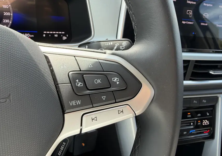 Gros plan sur le volant multifonction cuir du Volkswagen T-Roc gris, avec commandes intégrées et tableau de bord digital en arrière-plan.