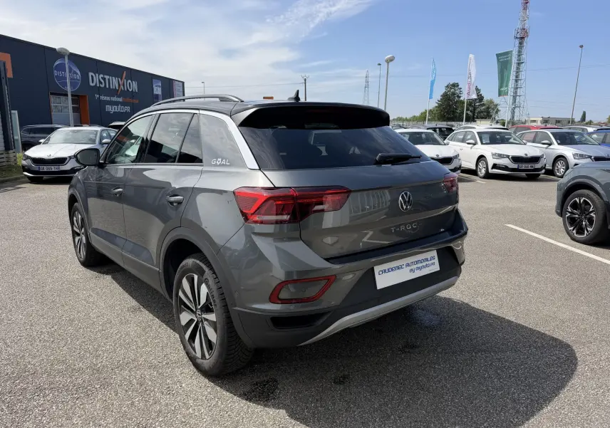 Vue 3/4 arrière droite d'un Volkswagen T-Roc gris avec barres de toit noires, sur parking extérieur ensoleillé.