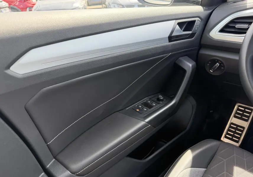 Vue intérieure côté conducteur du Volkswagen T-Roc 2025, montrant la portière noire avec surpiqûres blanches et commandes électriques.