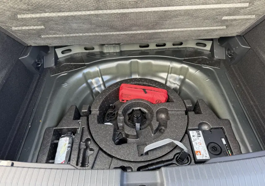 Vue du coffre ouvert d'un Volkswagen T-Roc gris, montrant le rangement sous plancher avec kit de réparation et trousse de secours rouge.