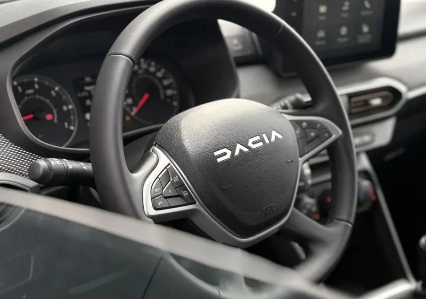 Vue rapprochée du volant noir Dacia avec commandes intégrées et tableau de bord numérique en arrière-plan.