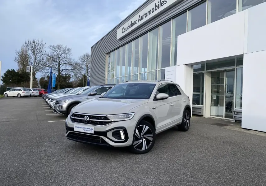 Volkswagen T-Roc gris en 3/4 avant droit, avec jantes alliage noires et calandre R-Line visible devant un concessionnaire.