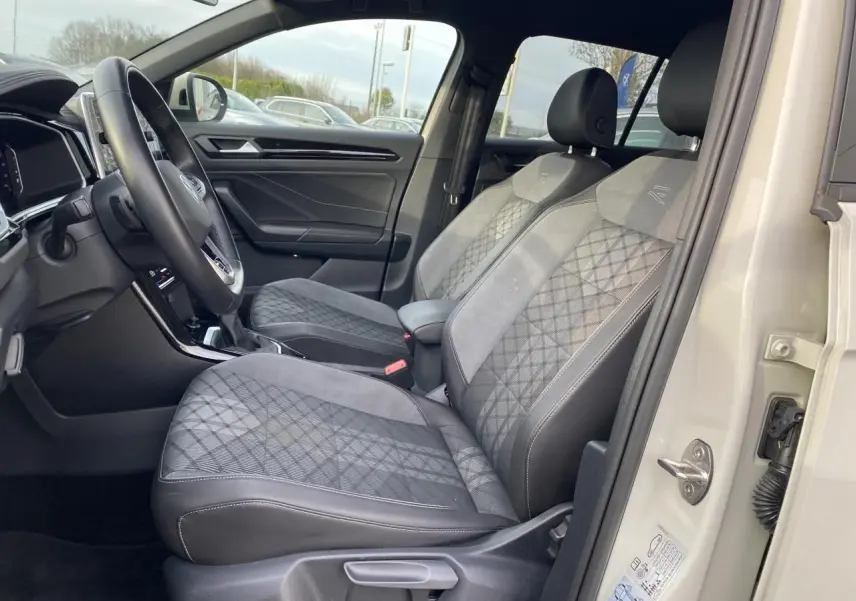 Vue intérieure côté conducteur du Volkswagen T-Roc 2024, sièges gris à motifs et volant multifonction en cuir.