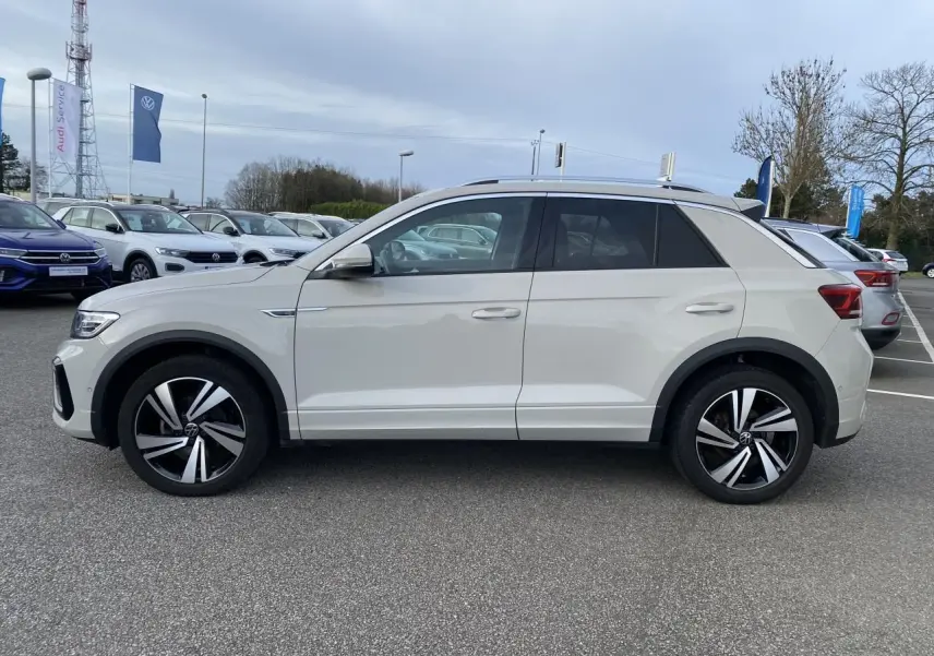 Vue de profil côté gauche d'un Volkswagen T-Roc gris clair avec jantes alliage bicolores sur un parking extérieur.