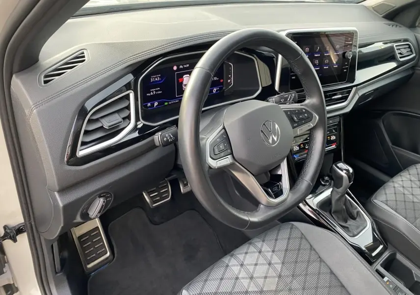 Vue intérieure côté conducteur du Volkswagen T-Roc 2024, volant cuir noir, tableau de bord digital et sièges tissu gris à motif.
