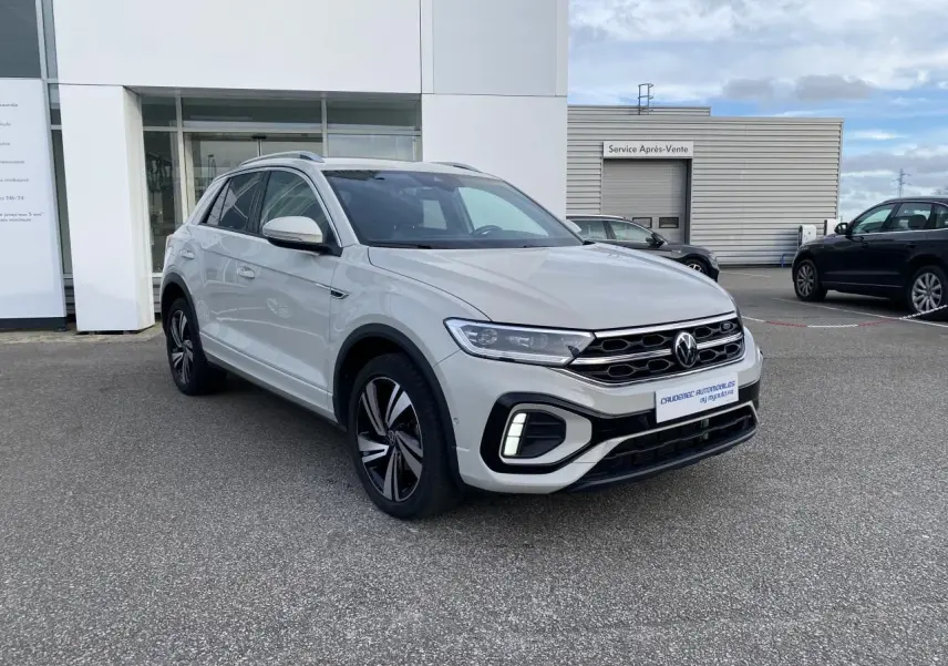 Volkswagen T-Roc gris clair en 3/4 avant droit, avec jantes alliage et calandre chromée visible.
