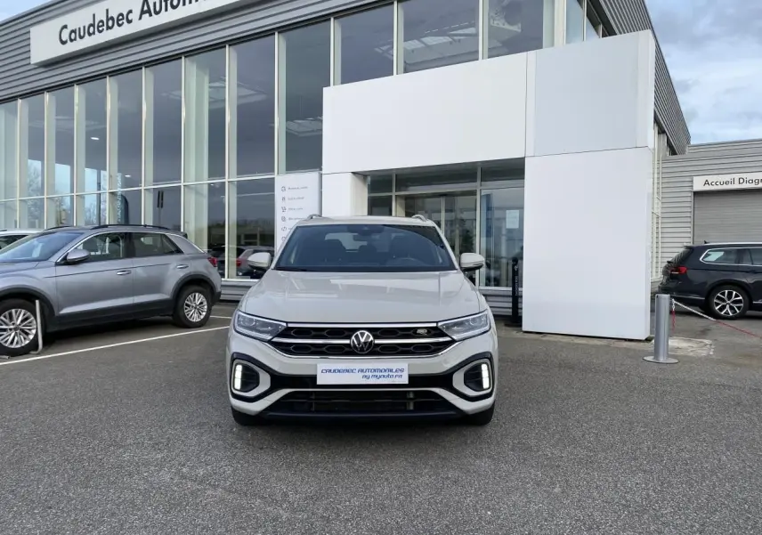 Volkswagen T-Roc gris vue de face, avec calandre noire et feux LED, stationné devant un showroom.