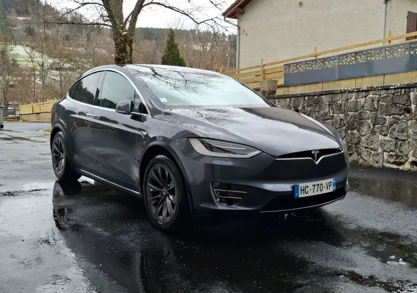 Tesla Model X 100D gris foncé vue 3/4 avant droit sur sol mouillé avec jantes alliage noires.