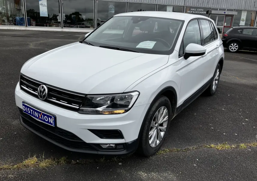 Volkswagen Tiguan blanc vu en 3/4 avant droit, avec calandre noire et feux allumés sur parking devant un hall d'exposition.