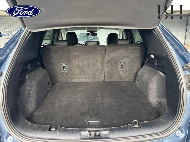 Vue intérieure du coffre ouvert d'un Ford Kuga bleu azur métallisé, montrant l'espace de chargement et la banquette arrière en tissu noir.