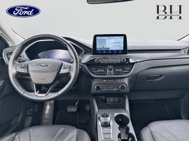 Vue intérieure avant du Ford Kuga 2023 avec tableau de bord noir, écran tactile central et volant cuir multifonction.