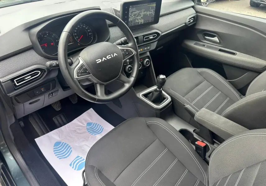 Vue intérieure côté conducteur du Dacia Jogger 2025, volant noir, tableau de bord avec écran tactile et sièges tissu gris foncé.