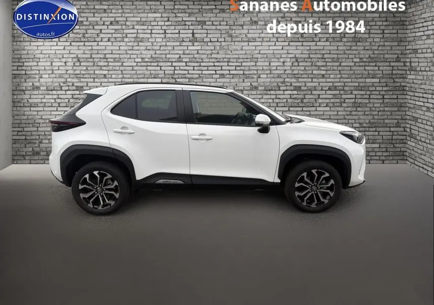 Profil côté gauche de la Toyota Yaris Cross Hybrid 130h blanche avec jantes alliage et protections noires sur fond de mur en briques.