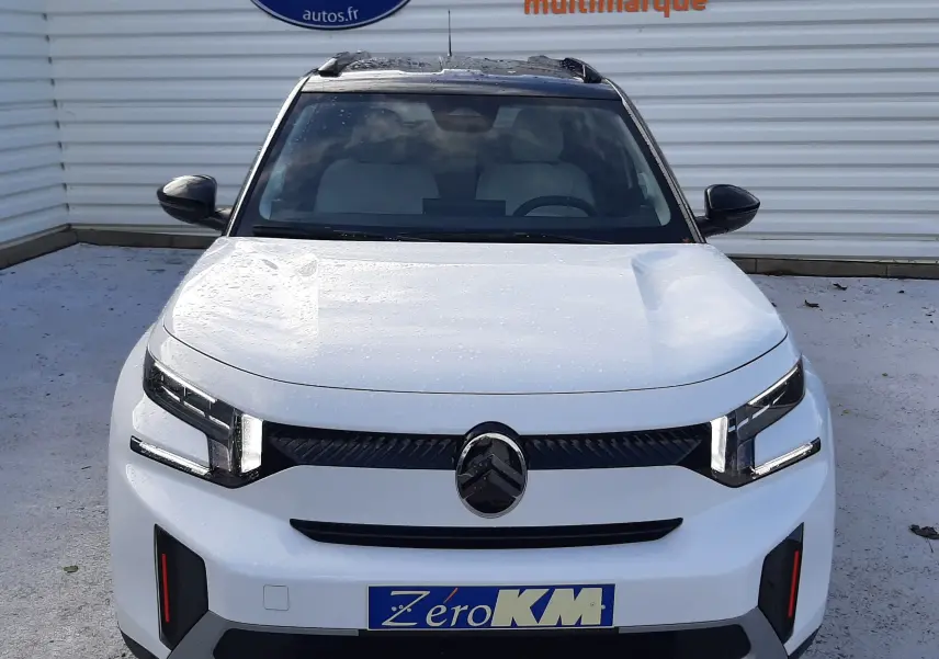 Vue 3/4 avant droite d'une Citroën C3 Aircross blanche avec toit noir, feux LED et jantes bi-ton, stationnée devant un mur blanc.