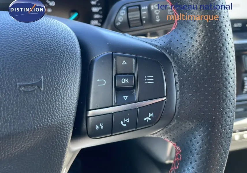 Gros plan sur le volant en cuir noir perforé avec surpiqûres rouges de la Ford Focus SW 2024, boutons de commande visibles.