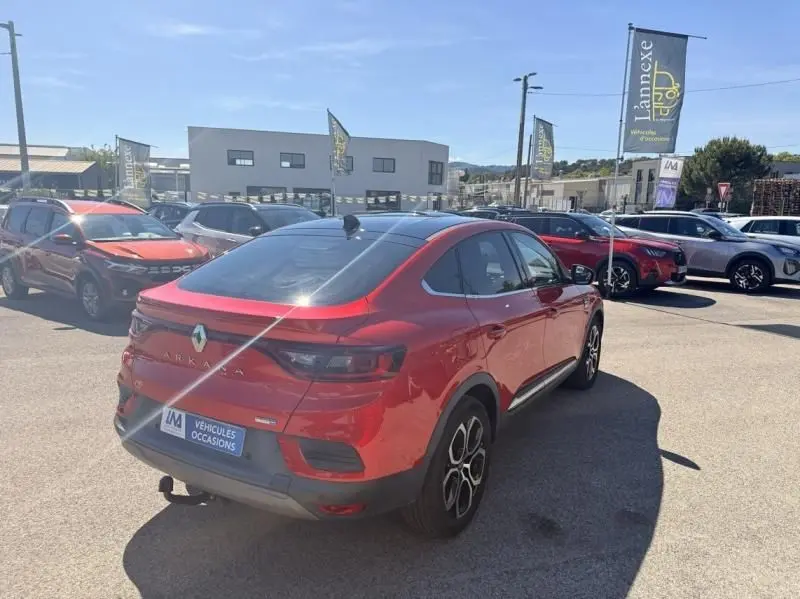 Vue 3/4 arrière droite d'un Renault Arkana 1.6 E-Tech rouge flamme avec toit noir, en concession sous un ciel dégagé.